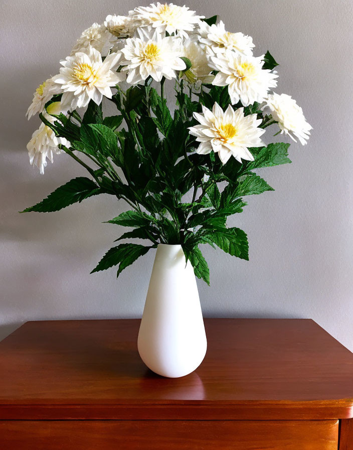 White chrysanthemums in white vase on wooden surface against neutral wall