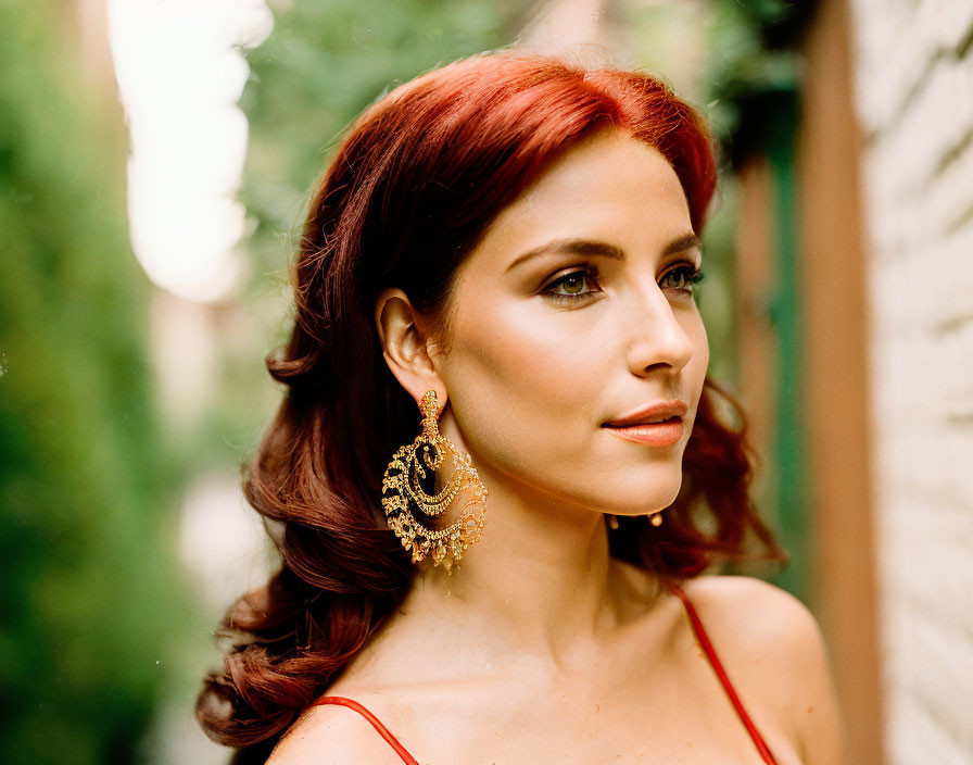 Red-haired woman in gold earrings wearing red dress and warm-toned makeup.