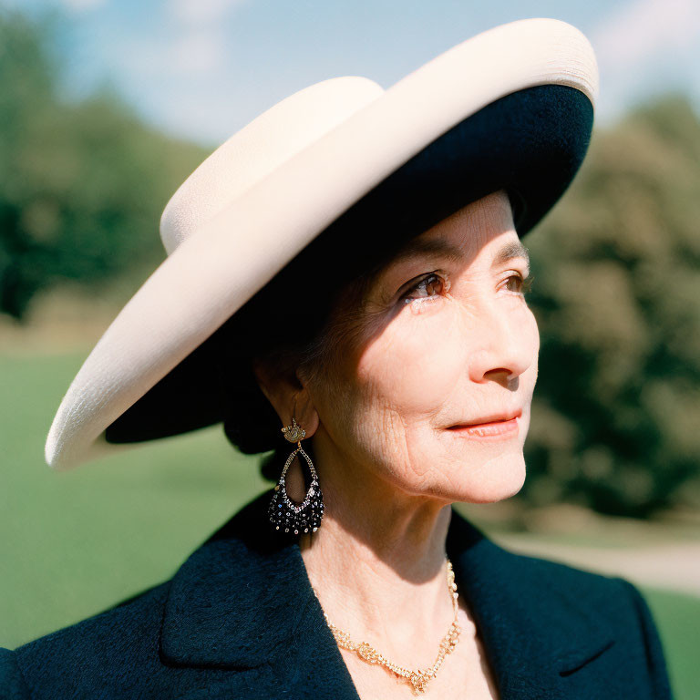 Senior woman in cream and black hat, dark blazer, and gold necklace against green backdrop