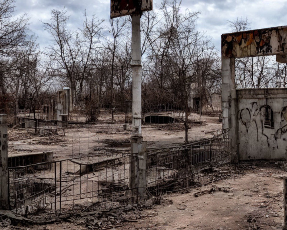 Abandoned area with bare trees, graffiti, and remnants under cloudy sky