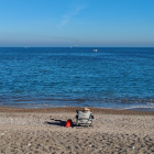 Person in towel on sandy beach gazes at blue sea with boats and red bucket.