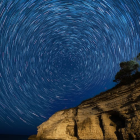 Night sky with circular star trails over mountain landscape and illuminated house.