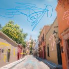 Colorful European street scene with cobblestone road, sidewalk cafes, and towering spire.