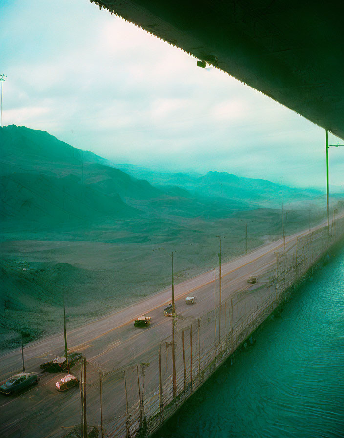 Vehicles on road under overpass with water, mountains, teal sky