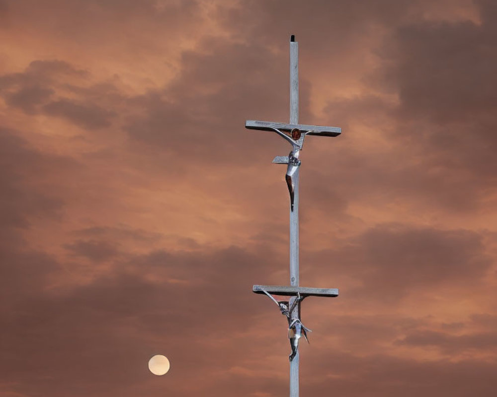 Metal figures on cross under orange dusk sky with white moon