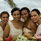 Four women in elegant dresses with floral crowns, one wearing angel wings, against serene landscape