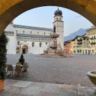 Couple embracing in cobblestone square with Christmas tree, fountain, and historical buildings.