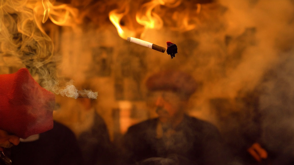 Person holding lit cigarette with smoke trail in red sleeve, blurred figures in smoky background