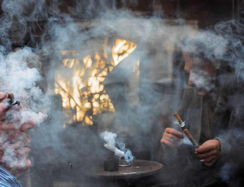 Two people in smoke with one exhaling and the other holding a smoldering object against blurred warm