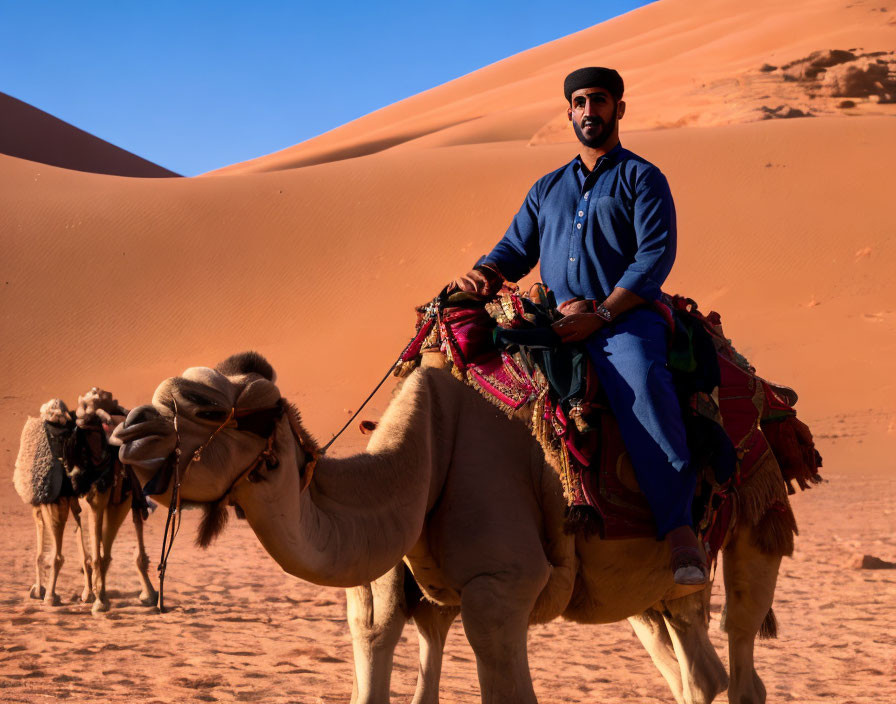 Traditional attired man riding camel in desert with sand dunes and more camels.