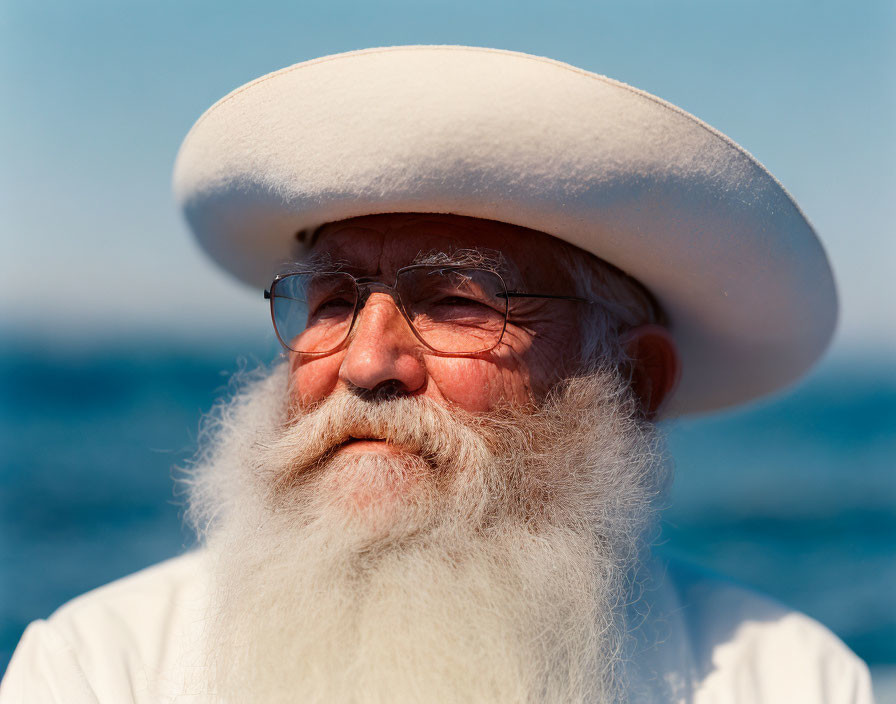 Elderly man with white beard, hat, glasses, by blue sea