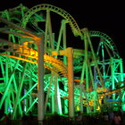 Night view of illuminated industrial complex with green and yellow piping, tanks, and worker silhouettes