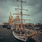 Tall ship with multiple masts in busy port with city skyline and dramatic sky