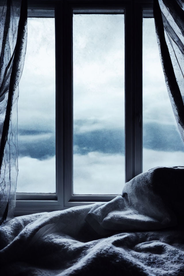 Cozy blanket by window with sheer curtains under cloudy sky