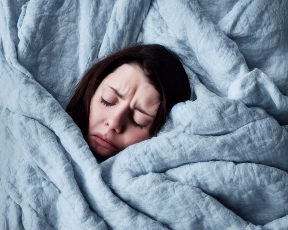 Person wrapped in blue-grey blanket with pained expression.