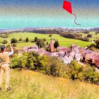 Man in vintage clothing flying red kite over picturesque village and green fields under sunny sky.