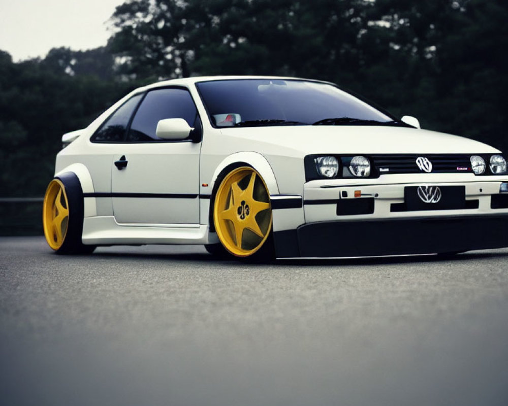 Customized white Volkswagen hatchback with yellow rims, body kit, and lowered suspension parked on a tree