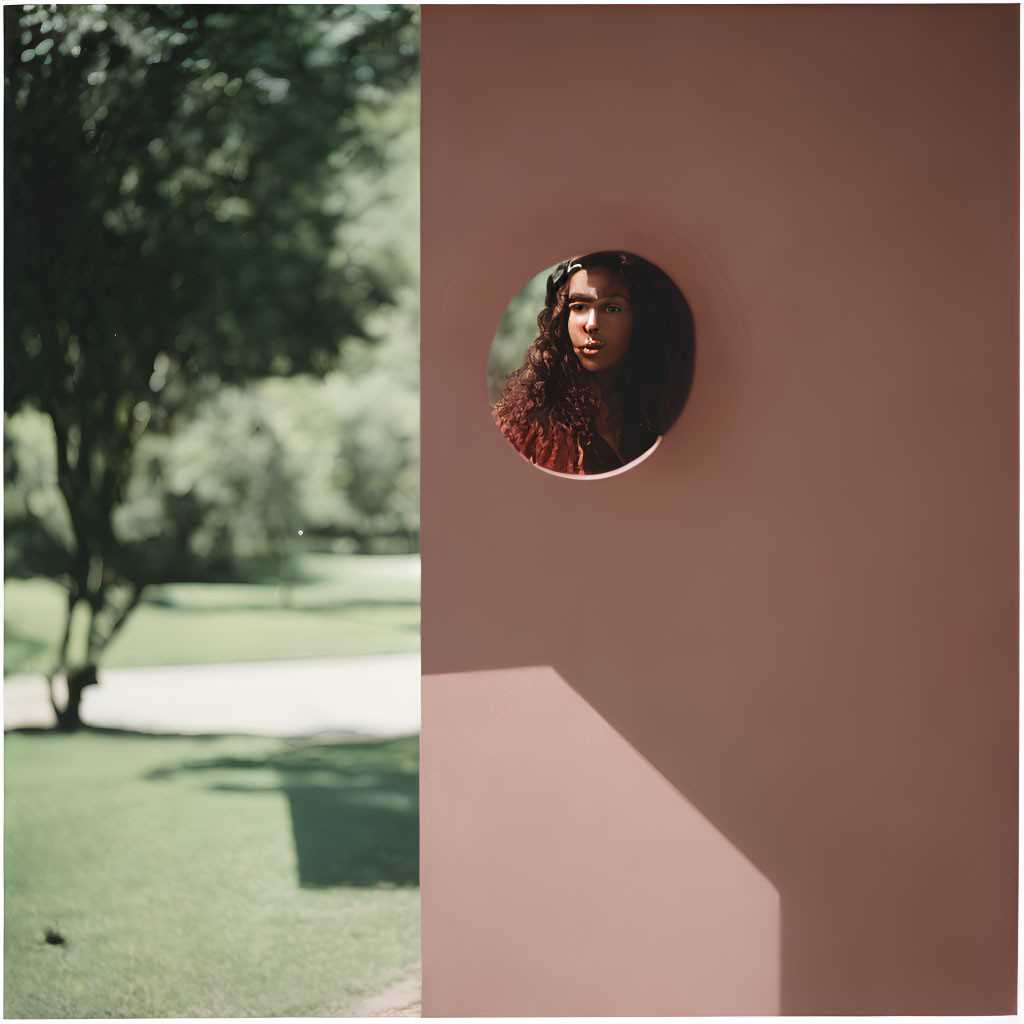 Woman looking through circular hole in pink wall with blurred green garden.