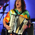 Long Curly-Haired Person in Robe Holding Oversized Accordion Under Spotlight