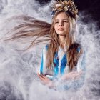 Young girl with long hair and floral crown in blue starry outfit among fluffy clouds