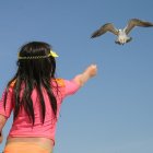 Young girl in colorful dress flying kite on sunny beach with distant child.