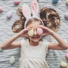 Girl in white dress with bunny ears holding colorful eggs on striped floor for Easter.