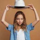 Pale-skinned girl with curly hair in surreal portrait wearing wide-brimmed hat and vintage pendant.