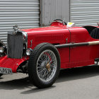 Vintage Red Convertible Car with Chrome Accents and Spoke Wheels Parked by Metal Gate