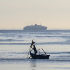 Person in small boat gazes at large ship on calm sea under gradient sky