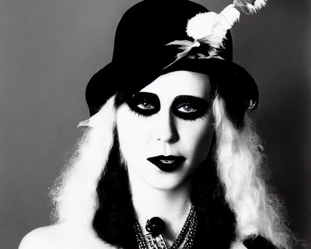 Monochrome image of person in dramatic makeup, feathered hat, layered necklaces, and curly hair