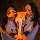Two Women in White and Gold Attire Holding Flame in Dark Setting