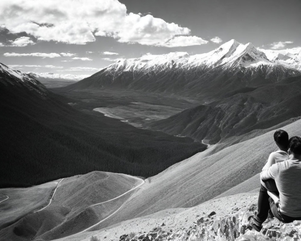 Two individuals on mountain edge with forested valleys and snowy peaks, black and white.