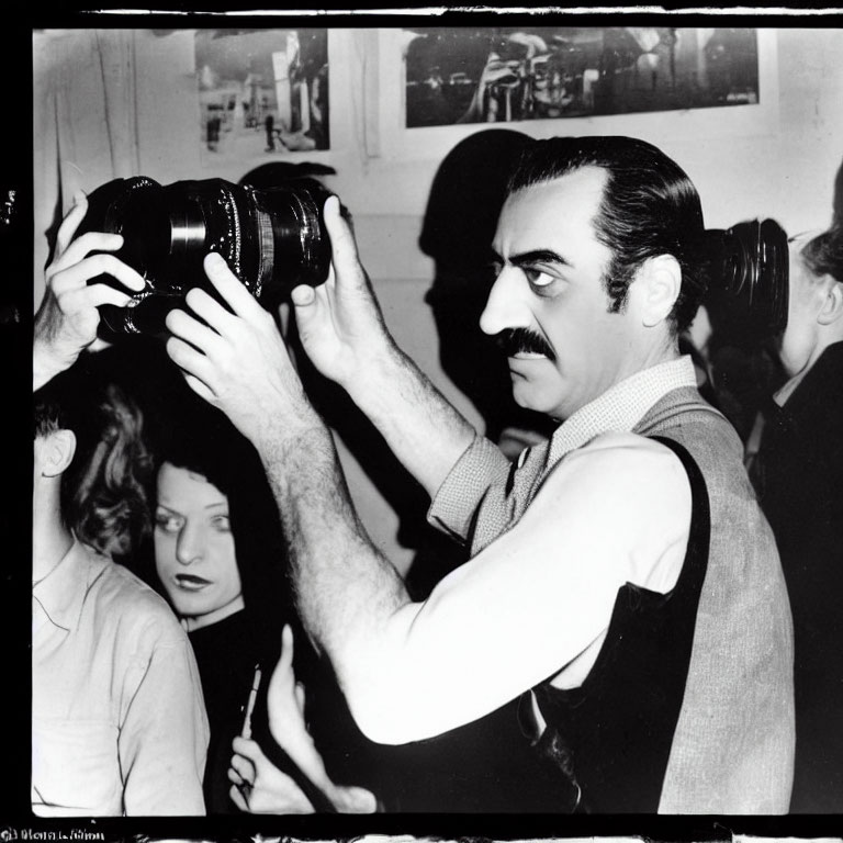 Monochrome photo: man with mustache holding camera, bystanders in background