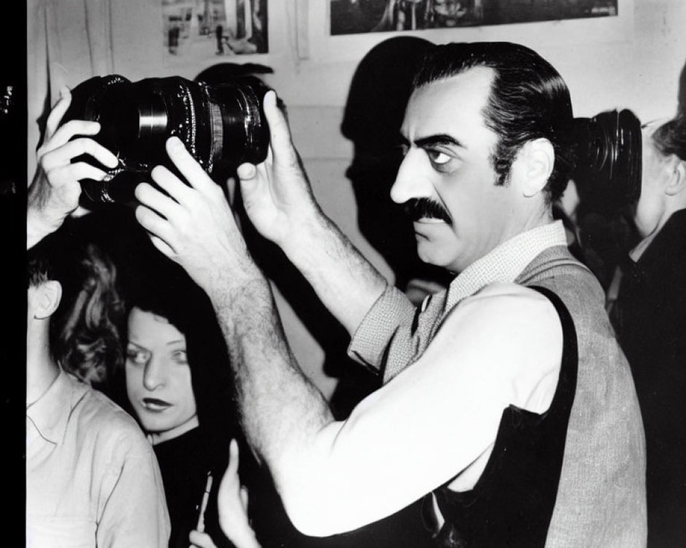 Monochrome photo: man with mustache holding camera, bystanders in background