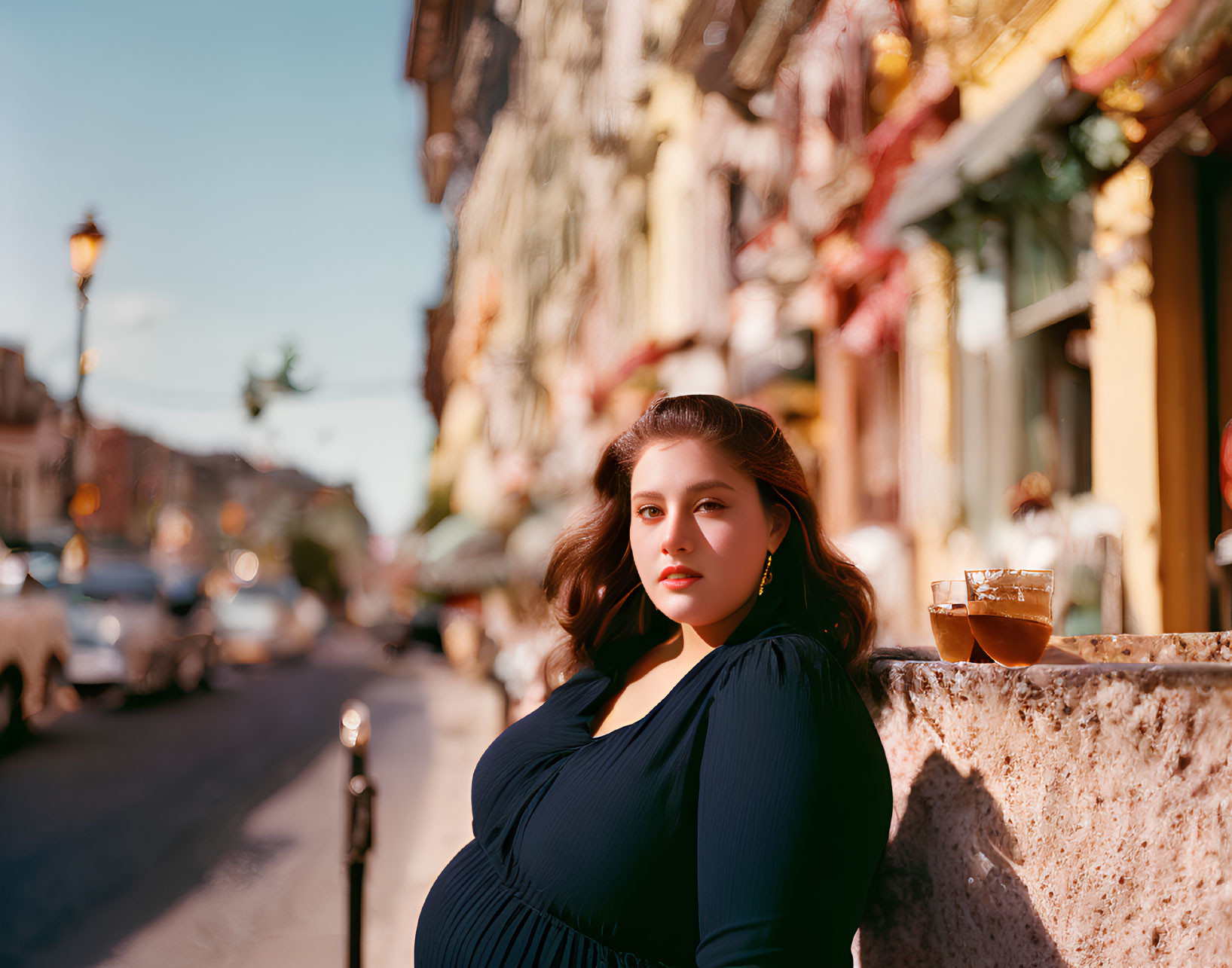 Woman in Blue Dress Leaning on Stone Ledge with Drink on Sunny City Street