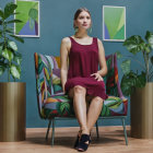 Stylish woman in burgundy dress on ornate chair with potted plants, teal background