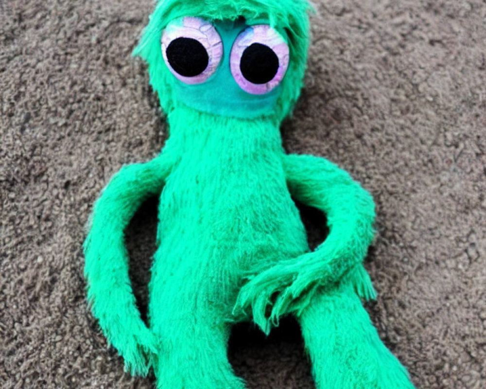 Green Plush Toy with Pink and White Eyes on Sand Surface