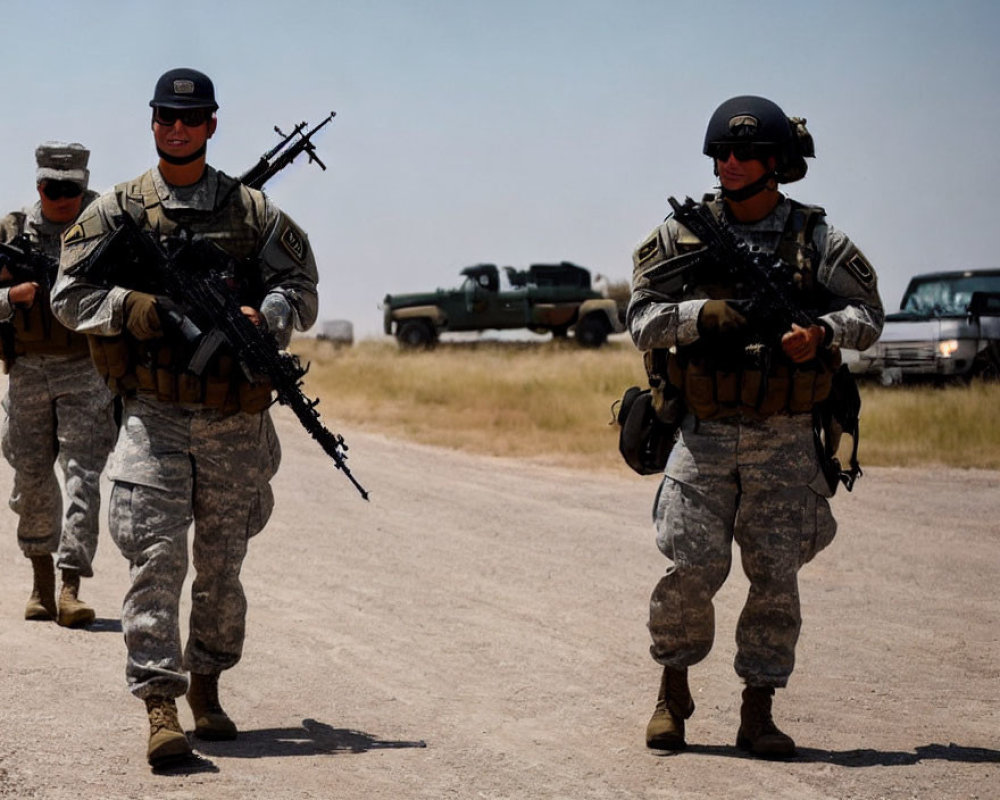 Military soldiers in camouflage walking on dirt path with rifles, vehicles in background.