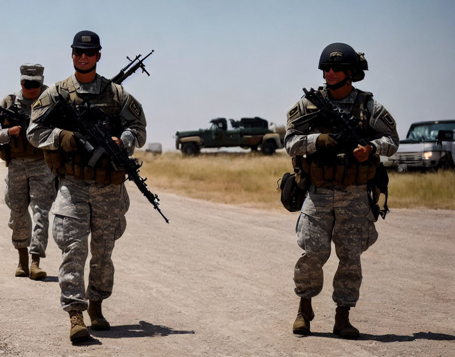 Military soldiers in camouflage walking on dirt path with rifles, vehicles in background.