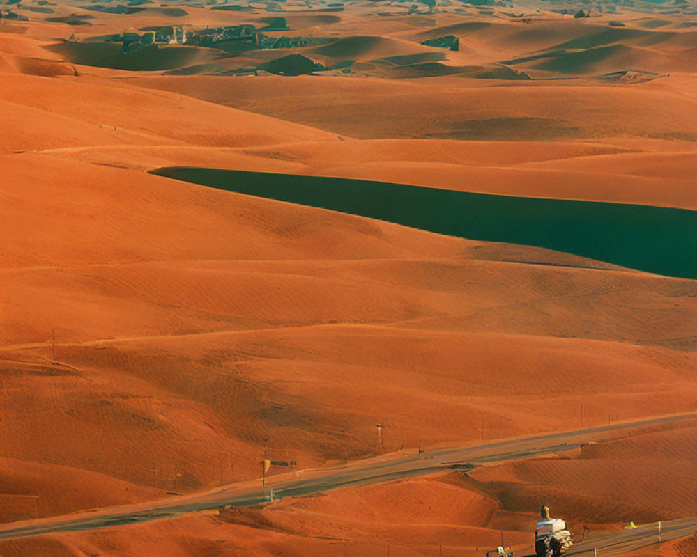 Desert road scene with truck, sand dunes, settlement, and setting sun