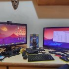 Dual monitor gamer setup with fantasy landscape wallpaper, colorful keyboard, decorative figurines, plants, and cozy