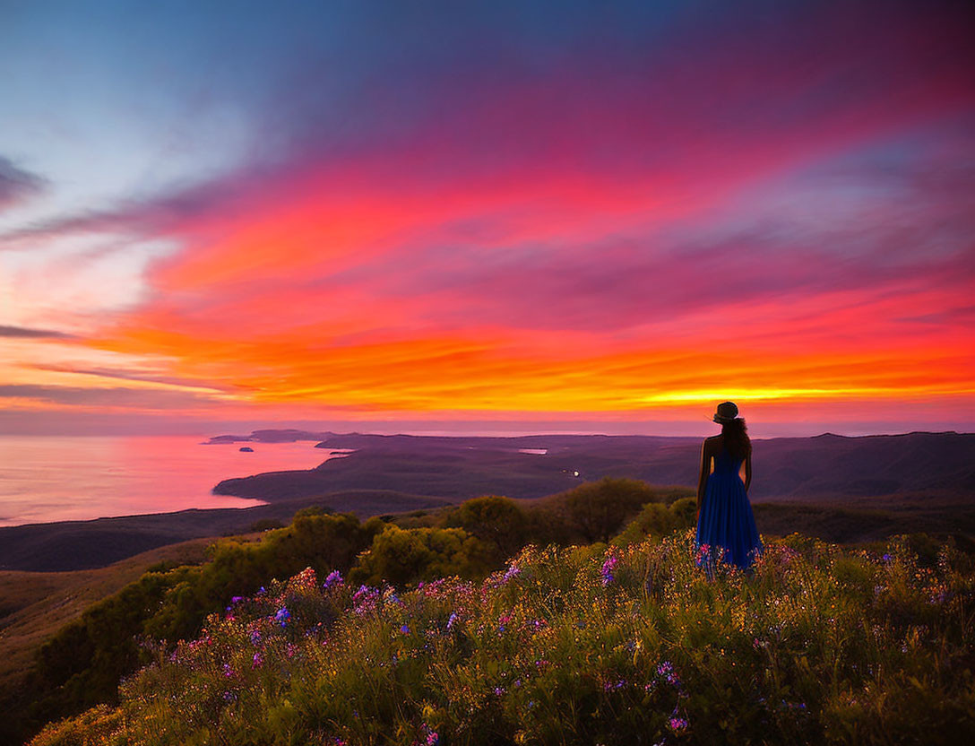 Person in Blue Dress Surrounded by Wildflowers at Vibrant Coastal Sunset