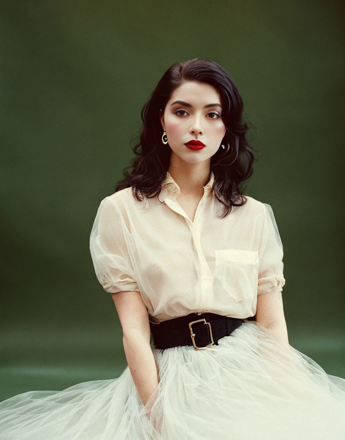 Dark-haired woman in sheer blouse and tulle skirt with red lipstick on green background