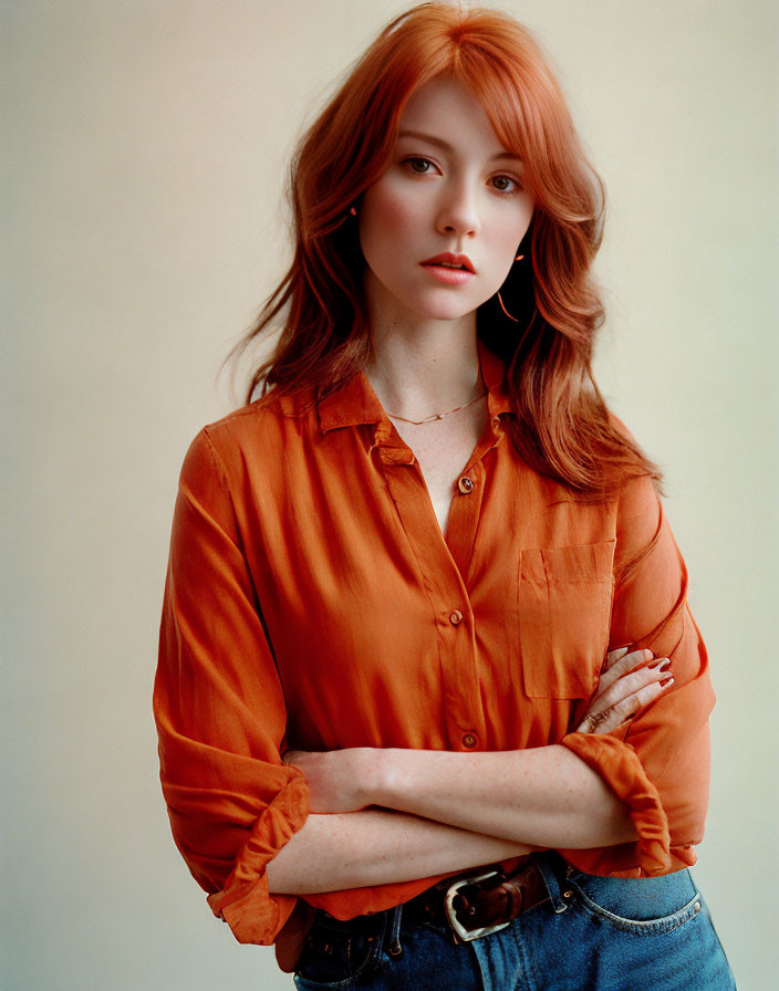 Red-haired person in orange shirt and blue jeans with crossed arms.