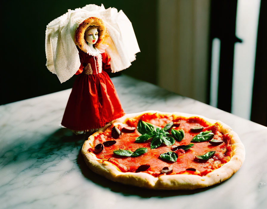 Vintage doll in red dress next to basil-topped pizza on marble surface