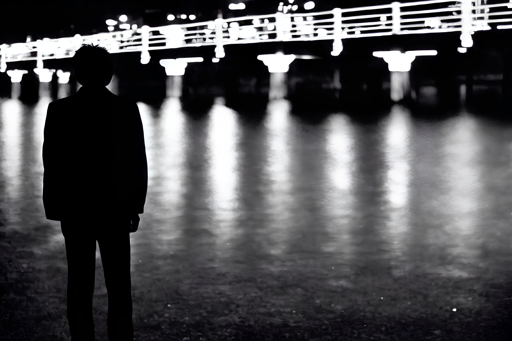 Silhouetted Figure by Water Reflecting Bridge Lights at Night