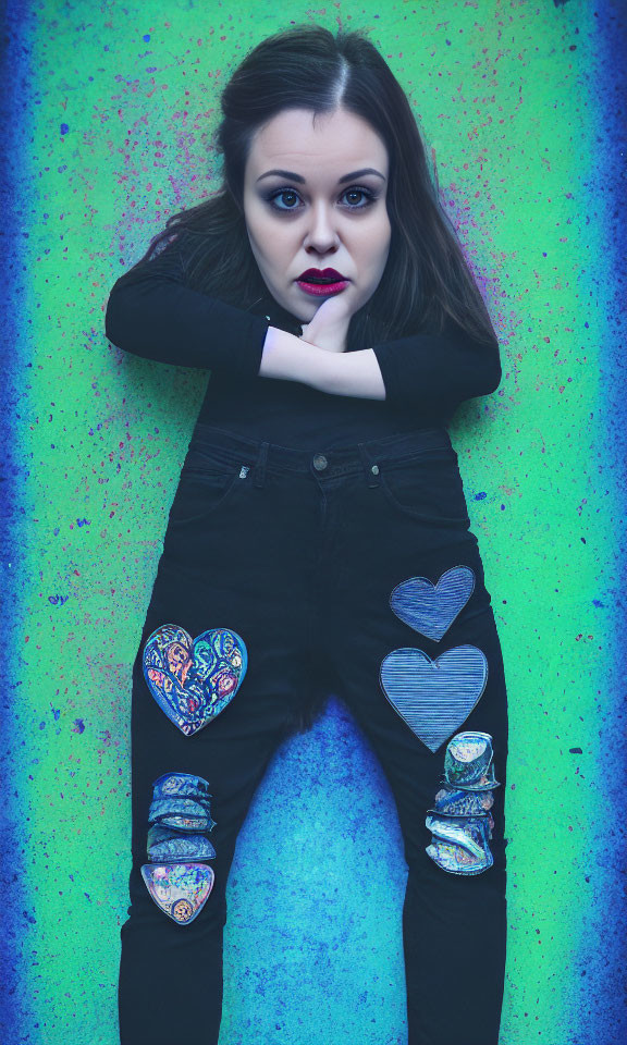 Dark-haired woman with bold makeup poses in black outfit with heart patches against colorful backdrop