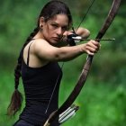 Woman with long braid aiming bow and arrow in navy outfit on green backdrop