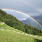 Colorful landscape with rainbow, pine forest, mountains, house, trees, and meadow.