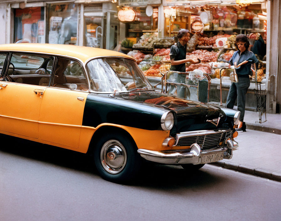 Vintage two-tone car on busy street with people and fruit stand.